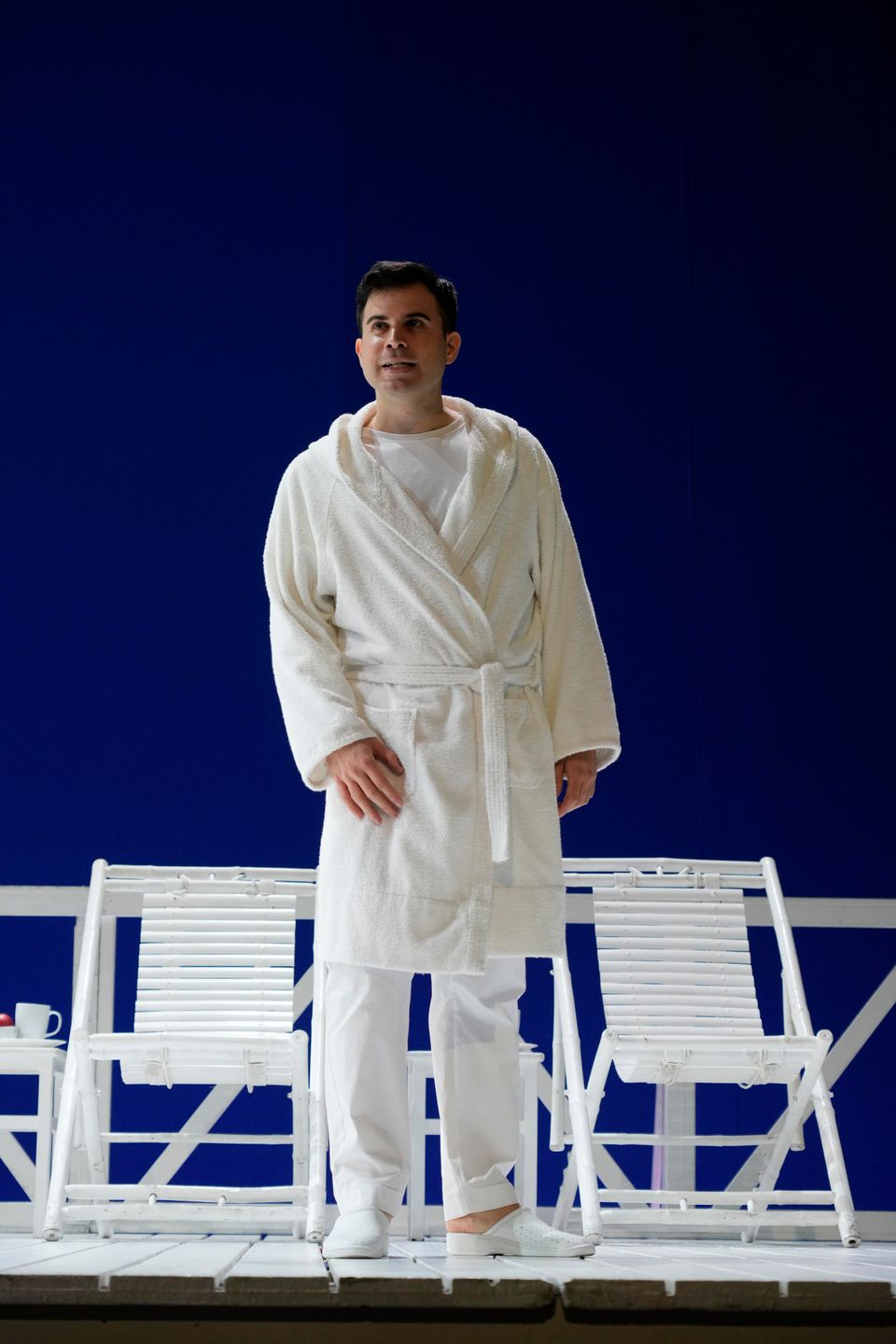 Enrico Basso in accappatoio bianco sul palco, davanti a due sedie a sdraio bianche e a un fondale blu.
