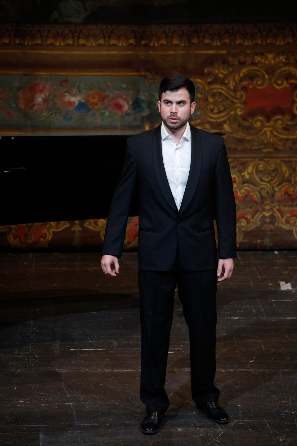 Enrico Basso in completo scuro e camicia bianca accanto a un pianoforte, davanti a una parete decorata con motivi floreali e dorati.